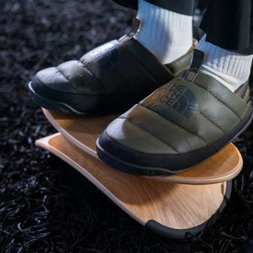 Person wearing insulated shoes with The North Face logo with a wood beflo strata footrest on a dark textured surface